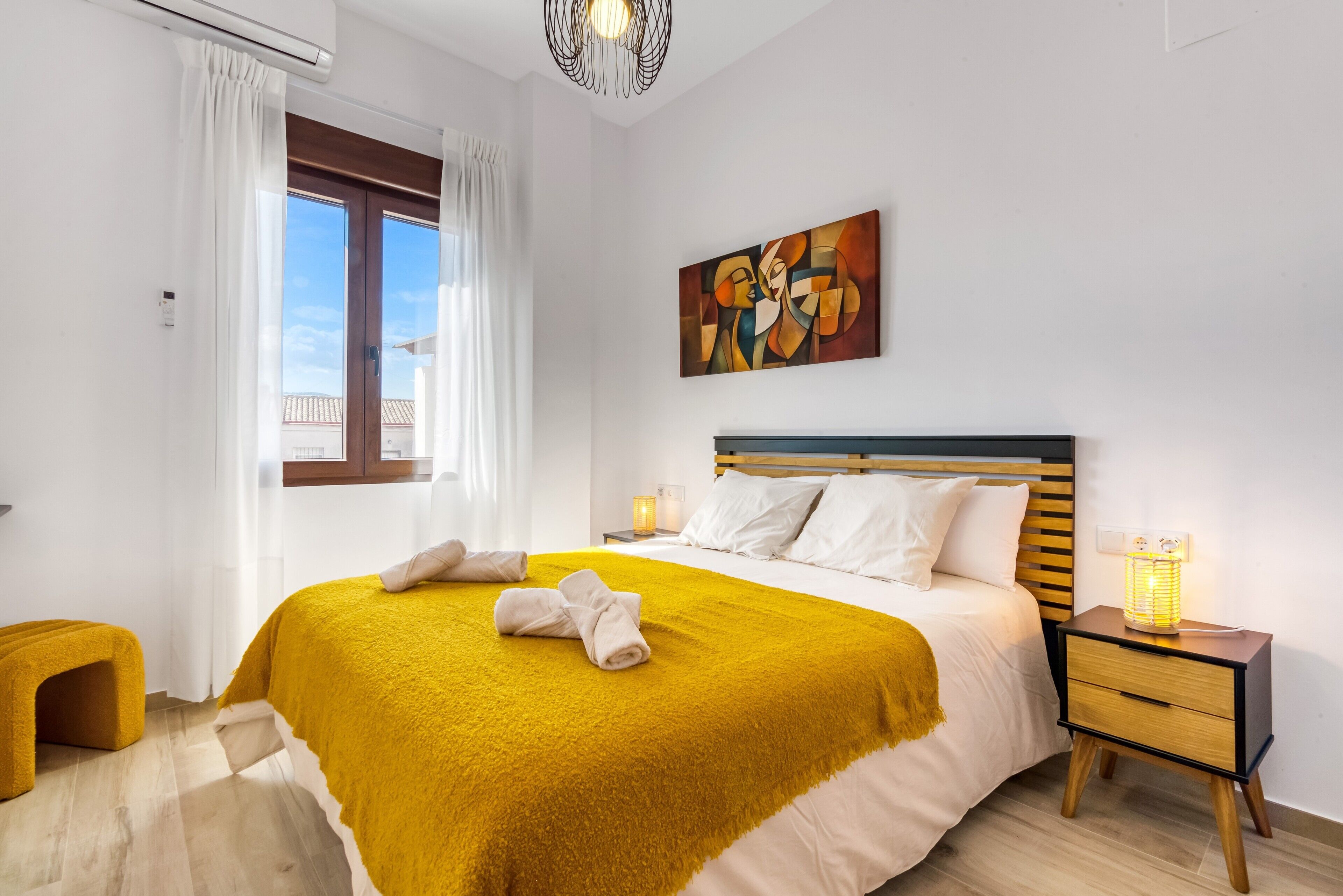 Ein Schlafzimmer mit Bett, Nachttisch, Lampe, Gemälde und einem Fenster mit Vorhängen.
