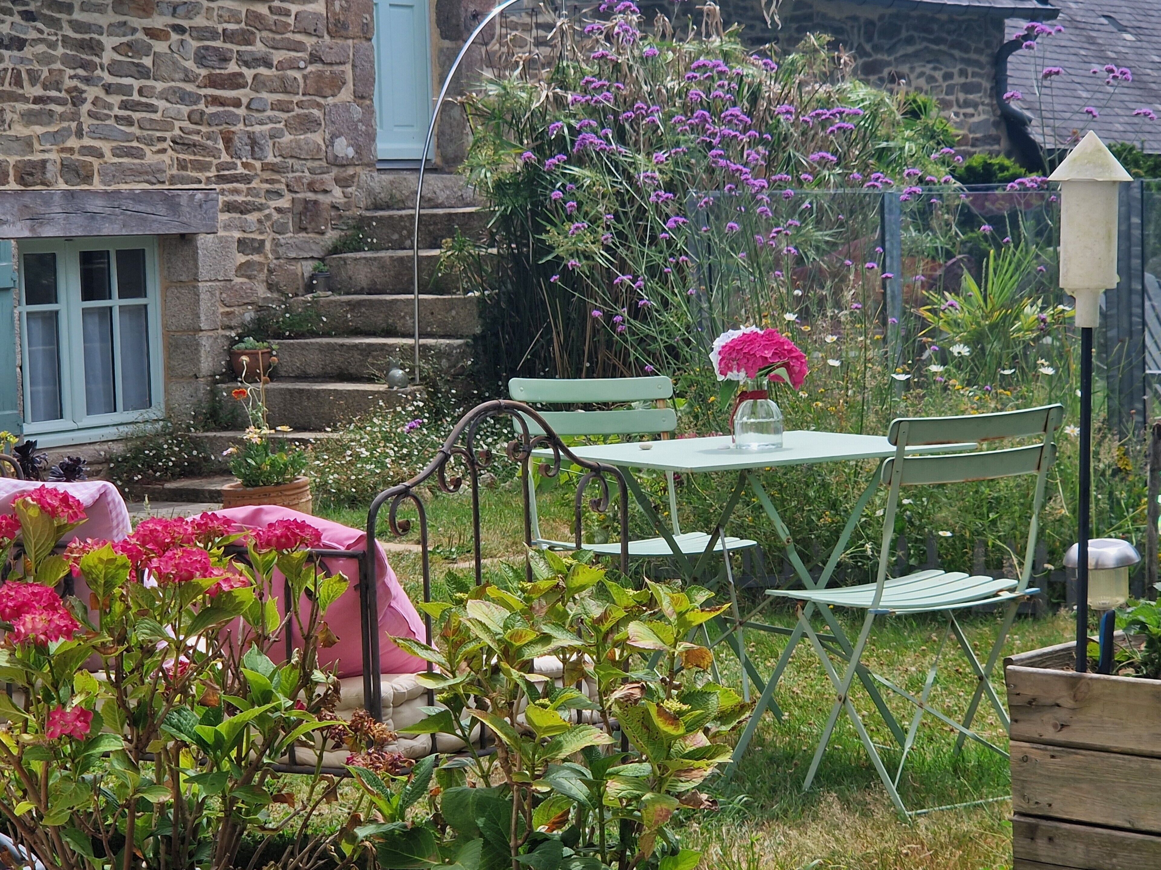 Een stenen gebouw met een trap, een tuin met diverse planten en bloemen, en tuinmeubilair, waaronder een tafel en stoelen.