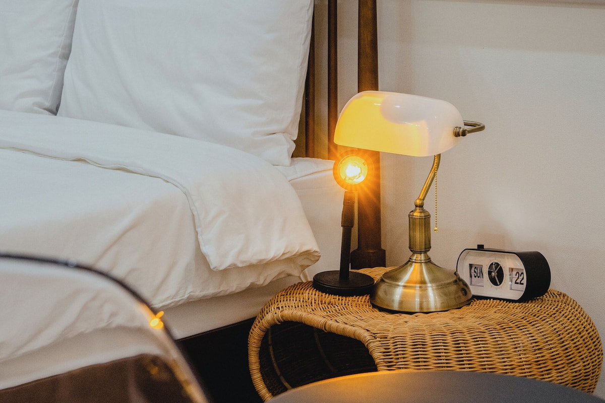 A neatly made bed with white bedding, a bedside lamp, a wicker side table, and a round table.