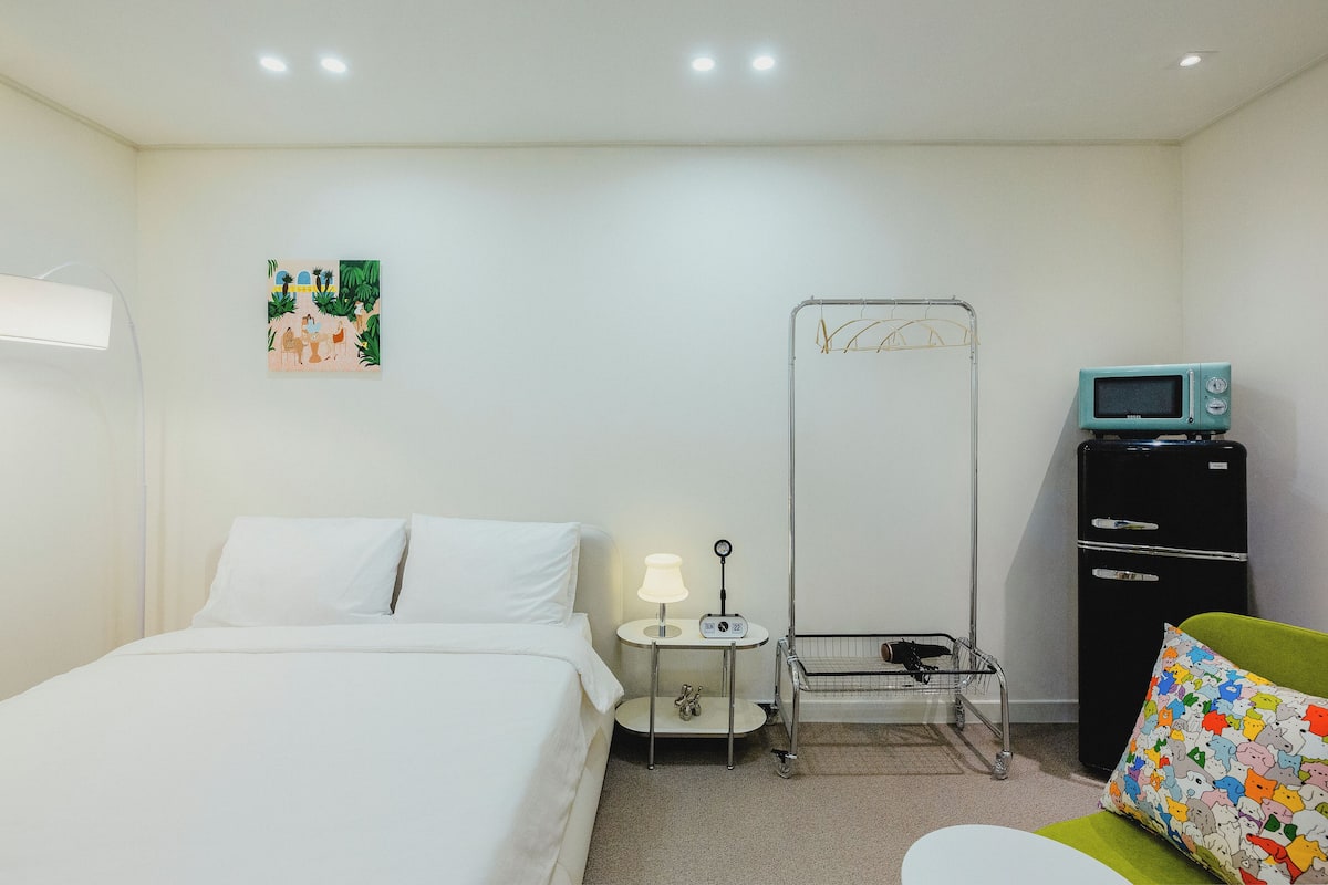 A hotel room with a bed, a small table with a lamp, a microwave, a refrigerator, and a colorful pillow.