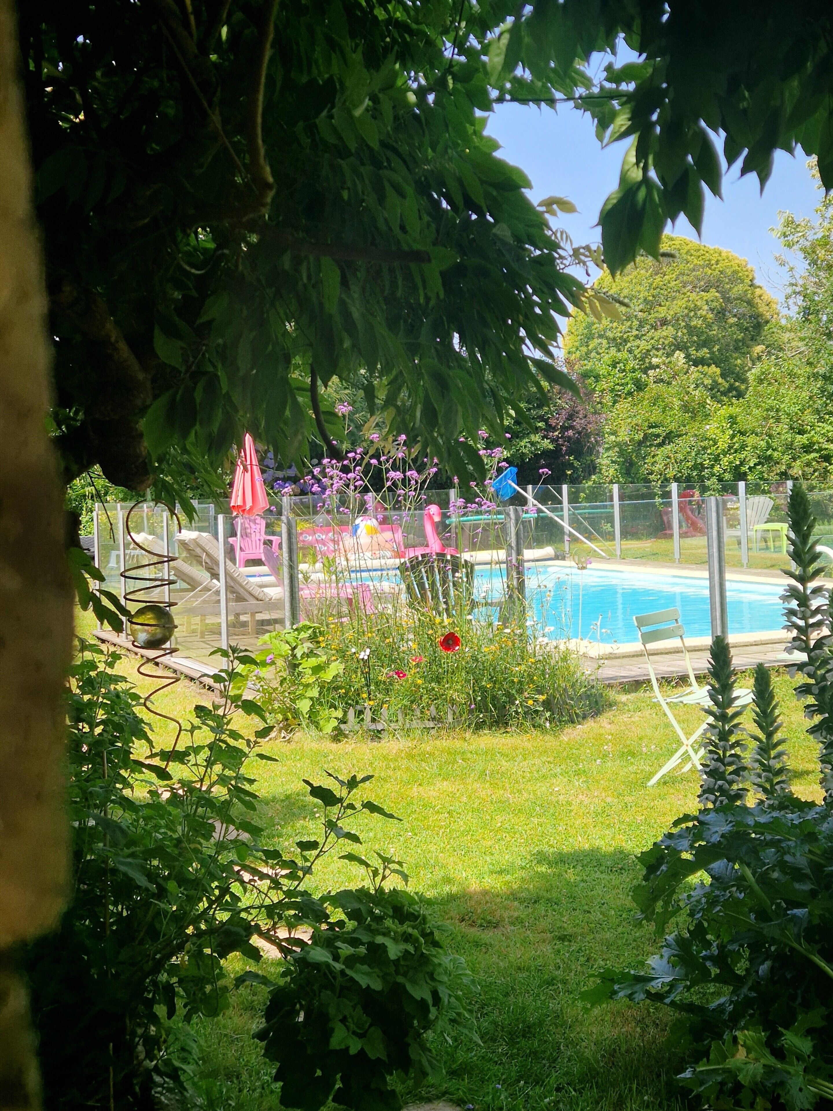 A swimming pool surrounded by a fenced garden with various plants and a playground area.