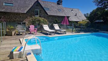 Une piscine avec des chaises longues et des parasols, un bâtiment en pierre avec un toit en tuiles et un ciel bleu azur.