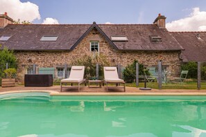 Une maison en pierre avec une piscine, deux chaises longues et une petite table.