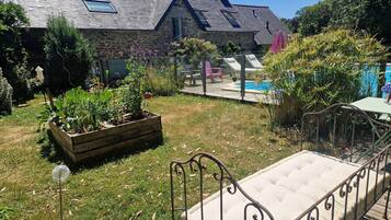 Una casa de piedra con techo inclinado, terraza de madera, jardín con bancales elevados y una banca de metal.