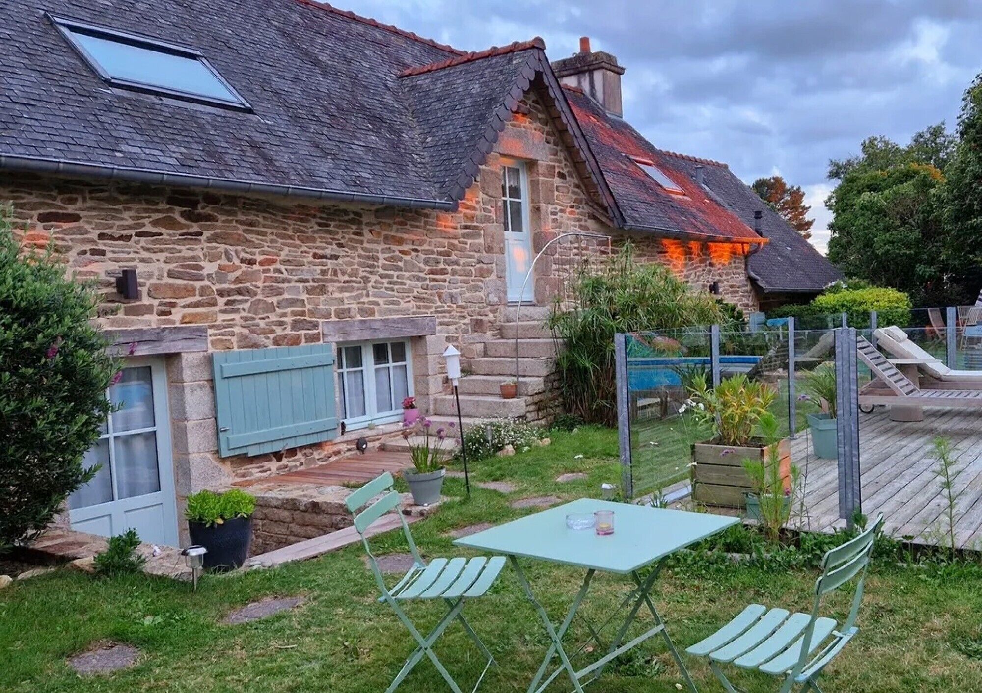 Een stenen huis met een schuin dak, een kleine tuin met een zwembad en terras.