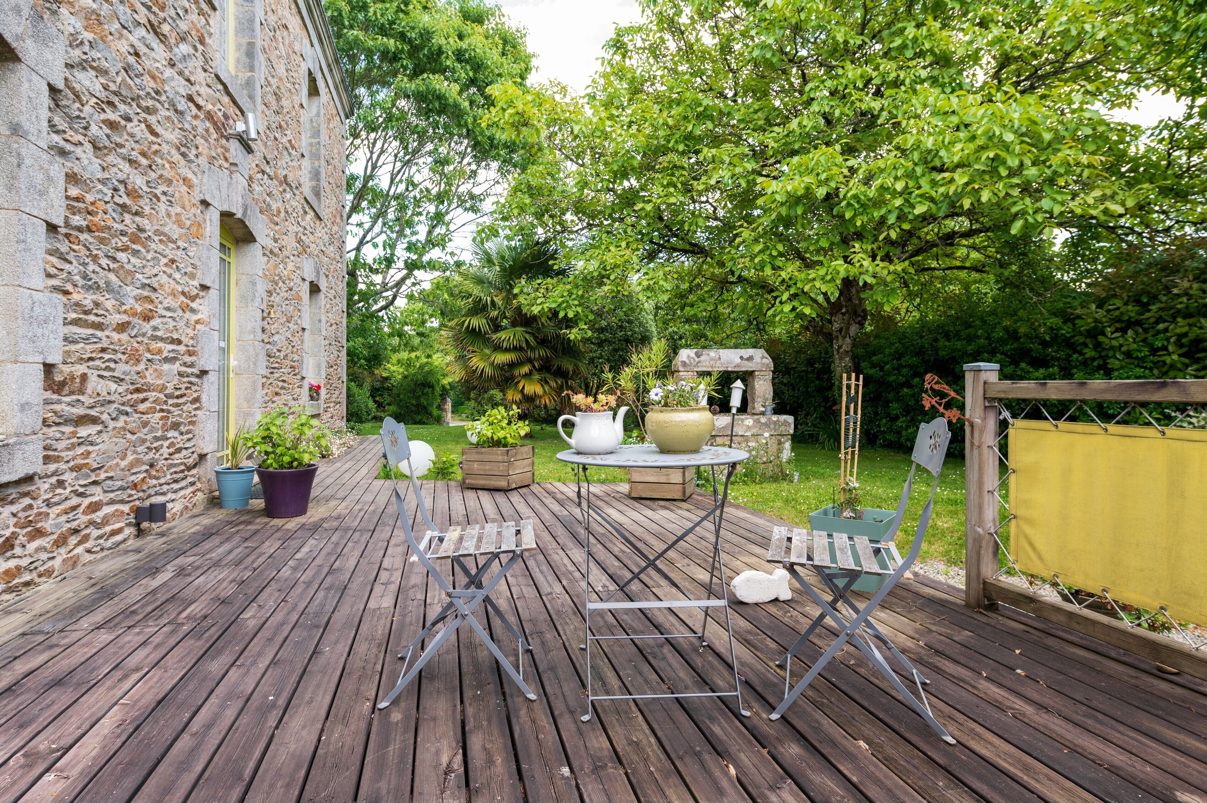 A wooden deck with outdoor furniture, a stone building, and a garden with trees and plants.