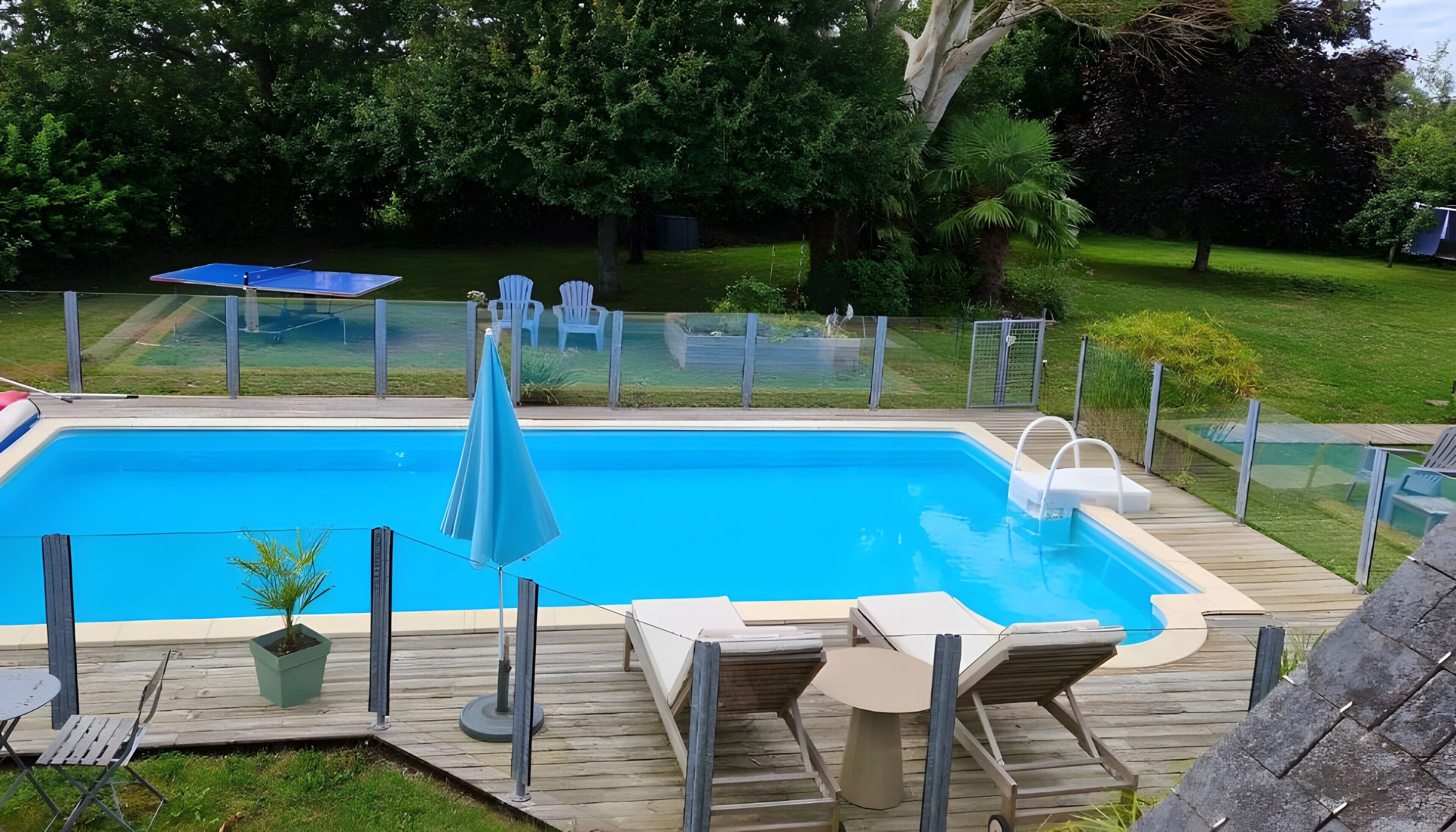 A rectangular swimming pool with a wooden deck, lounge chairs, and a small table.