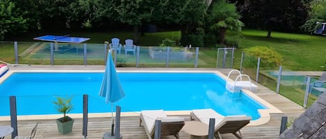 A rectangular swimming pool with a wooden deck, lounge chairs, and a small table.