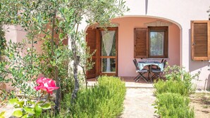 Un patio con una mesa y sillas, una puerta con ventanal, y un jardín con plantas verdes y un árbol.