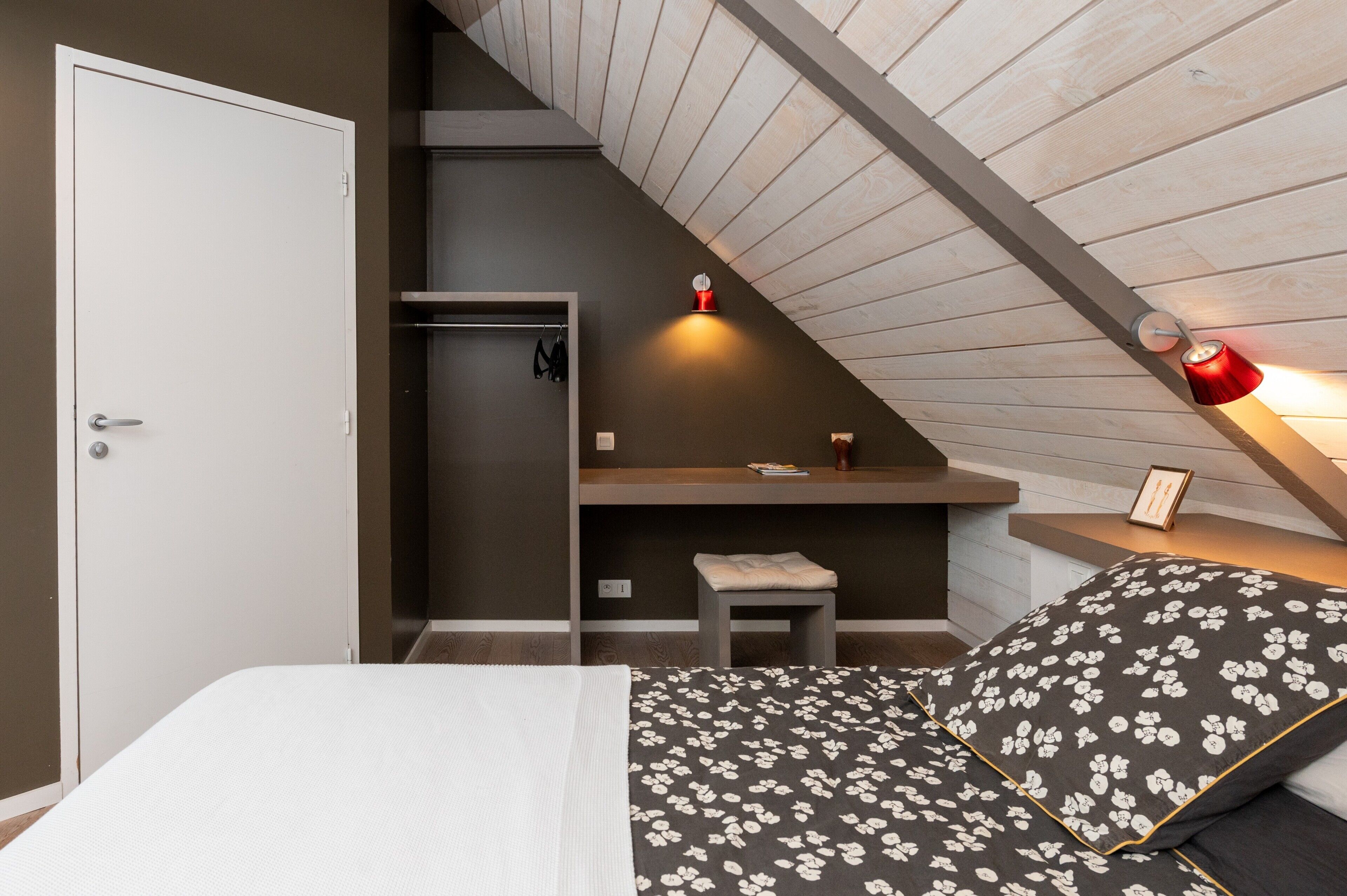 Une chambre avec un plafond en pente, un lit recouvert d’une couvre-lit à motifs floraux, une table de chevet avec une lampe rouge et un petit tabouret.