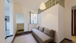 A compact living space with a sofa, a small window, a radiator, and a kitchen area visible through an open door.
