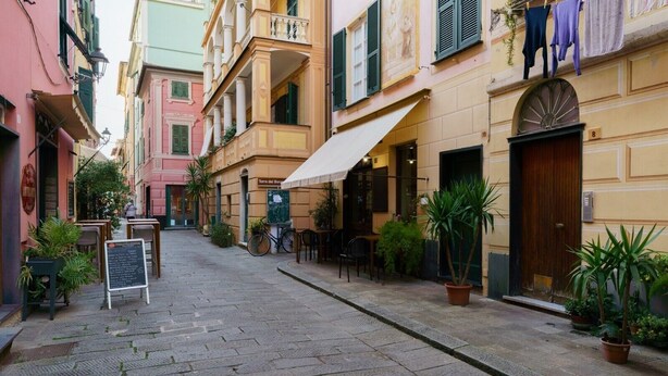 A narrow street with buildings on either side, outdoor seating, and potted plants.