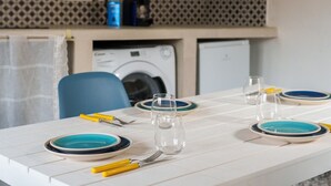 A dining table set with plates, glasses, and cutlery, a washing machine, and a blue chair in a room with a patterned wall and a curtain.