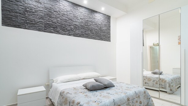 A bedroom with a bed, a nightstand, a mirror, and a textured wall. - Apartment 'Dafne Home' with Wi-Fi and Air Conditioning (Siracusa)