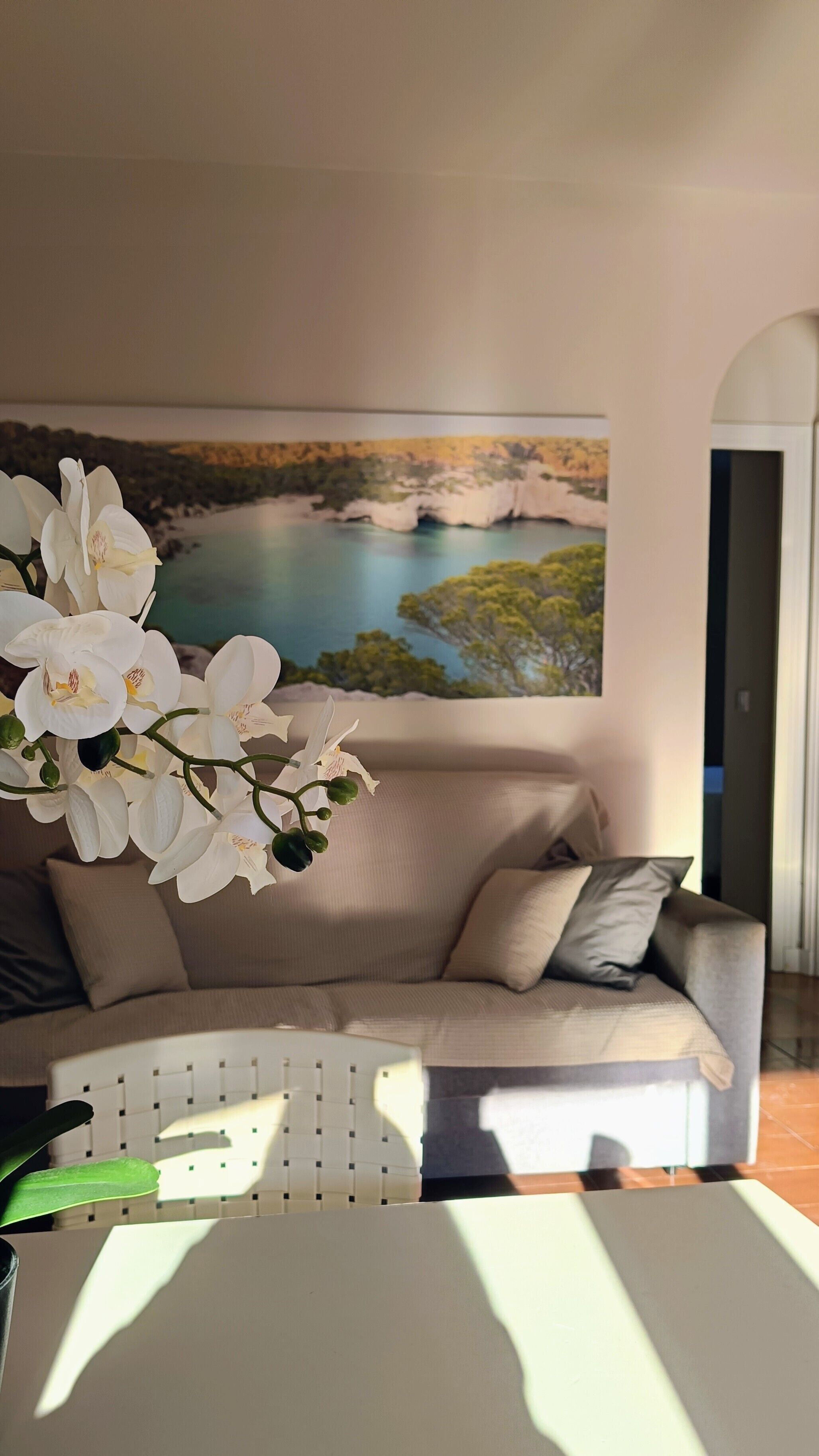 Una sala de estar con un sofá, un cuadro de un paisaje y orquídeas blancas.