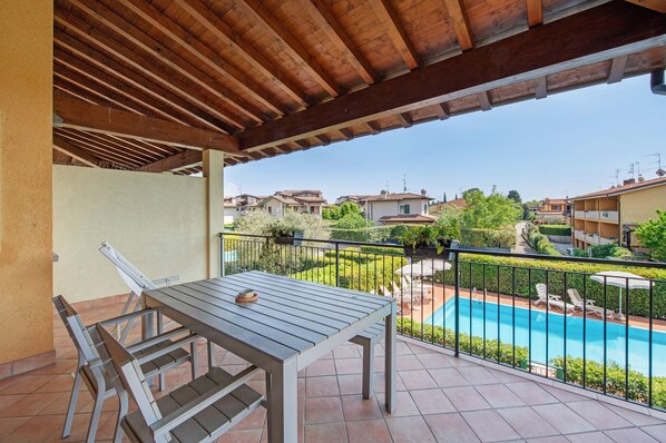 A covered outdoor area with a table and chairs, a pool, and a view of surrounding buildings.