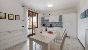 Cozinha moderna com armários brancos, uma mesa de jantar e cadeiras. Há uma janela com cortinas, vista para o exterior e piso revestido de azulejos.