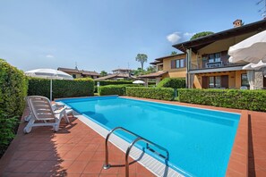 Una piscina con una escalera metálica, sillas de descanso y sombrillas.
