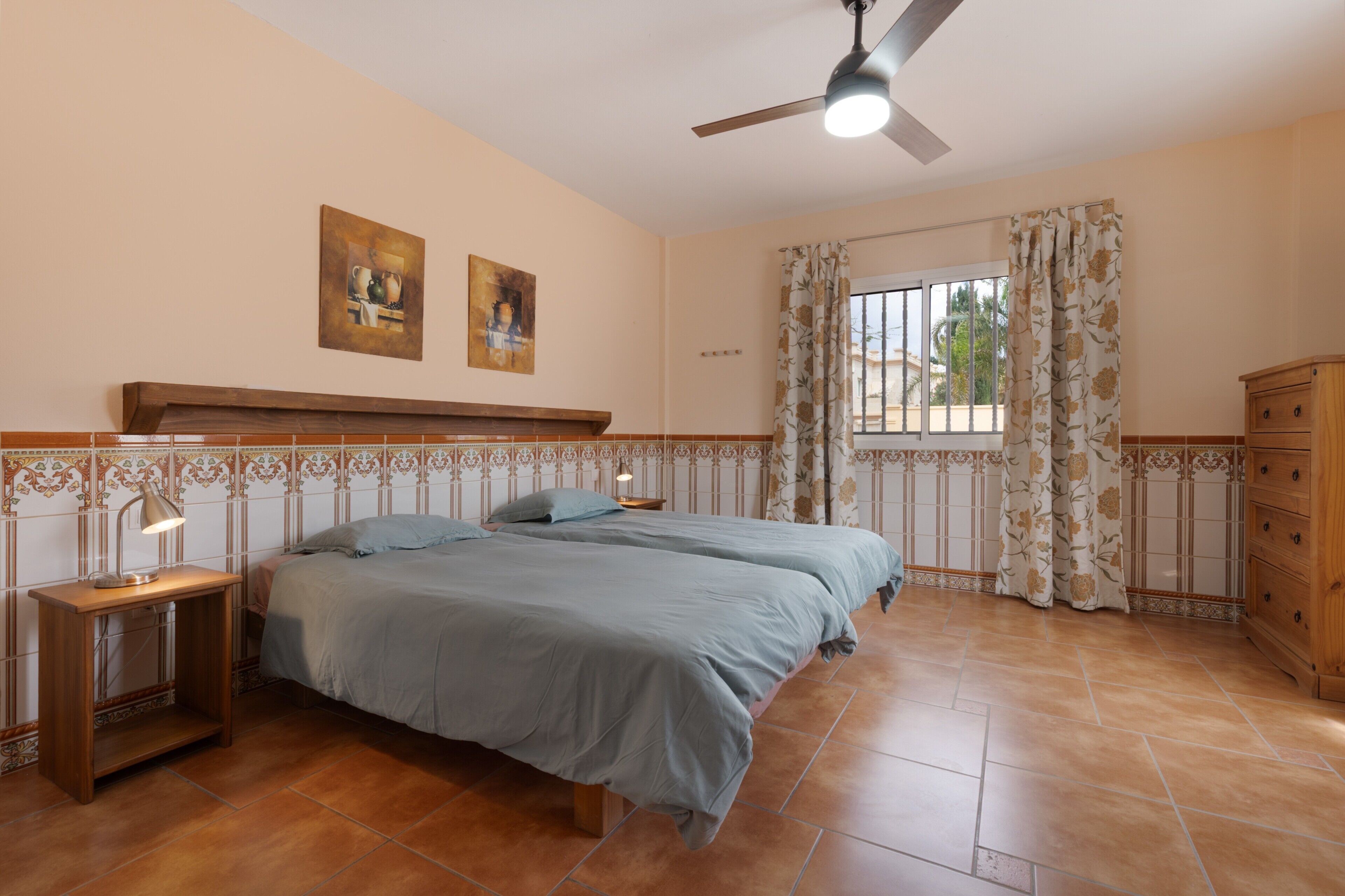 Un dormitorio con dos camas, ventilador de techo, ventana con cortinas y cómoda de madera.