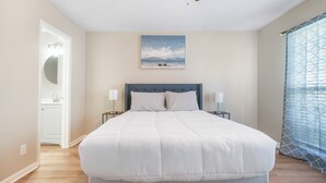 A bedroom with a large bed, a ceiling fan, a painting of a beach scene, and a window with patterned curtains.