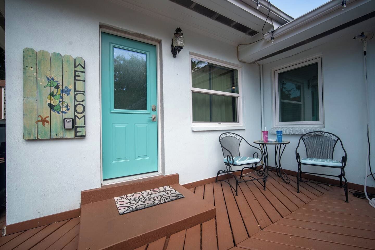 Une terrasse en bois avec une porte turquoise, deux fenêtres et deux chaises en métal.
