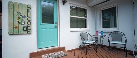 Una terraza de madera con una puerta turquesa, dos ventanas y dos sillas de metal.