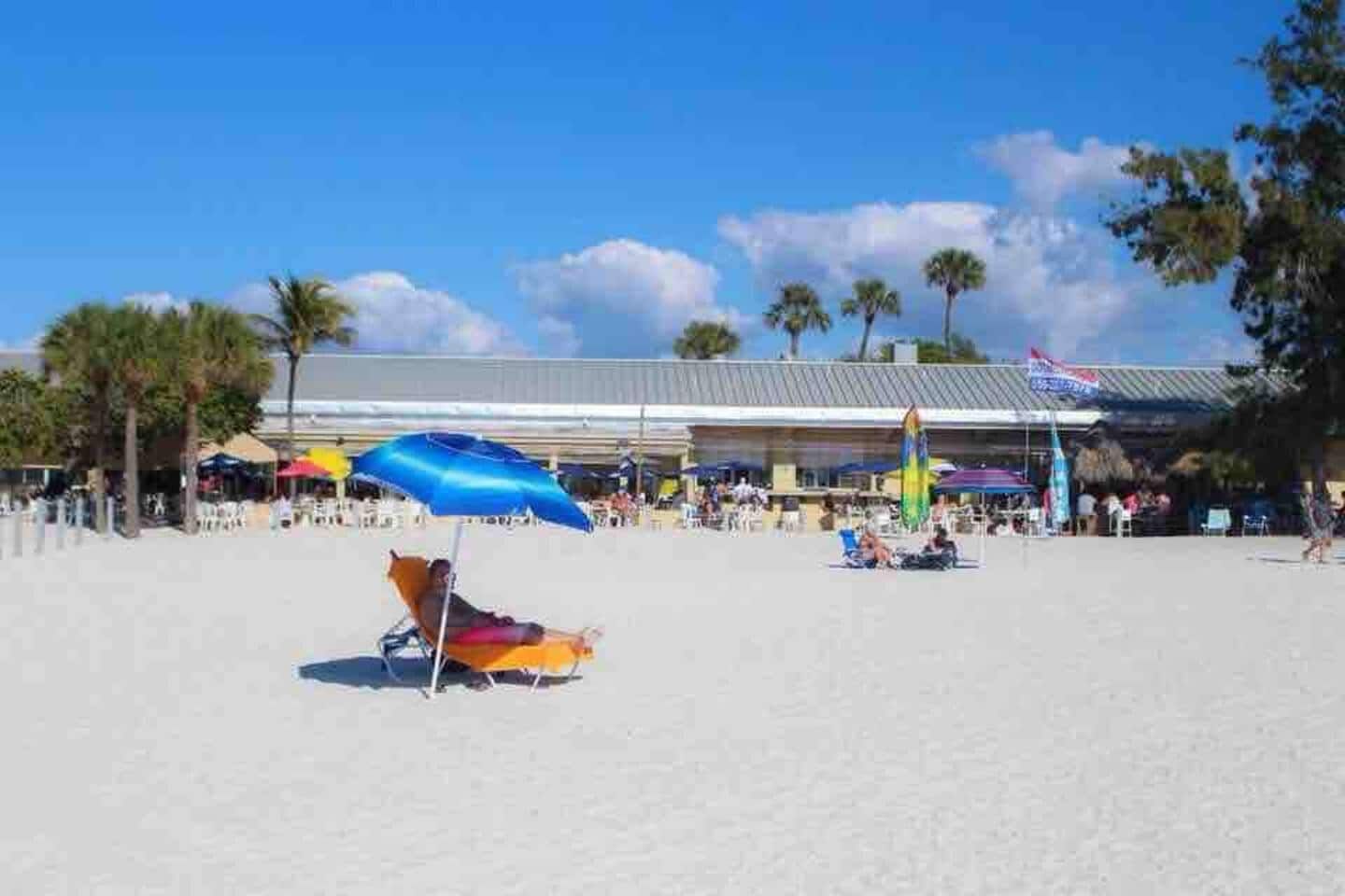 A beach with umbrellas, palm trees, and a building in the background.