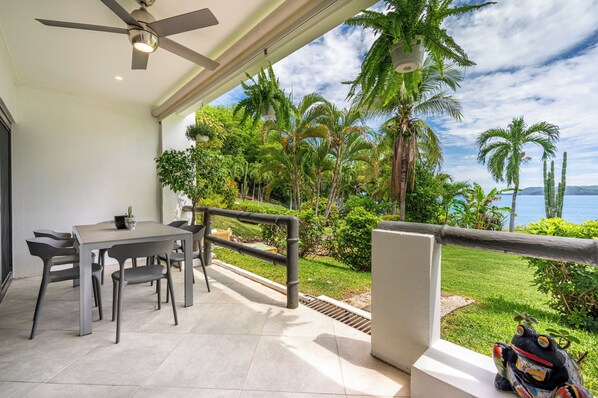 A balcony with a table and chairs, overlooking a lush garden and a body of water.