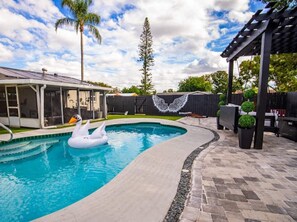 A swimming pool with a swan float, a covered patio area, and a fenced backyard with a mural. - Anna Maria Island Getaway | Pool + Game Room | IMG (Bradenton)