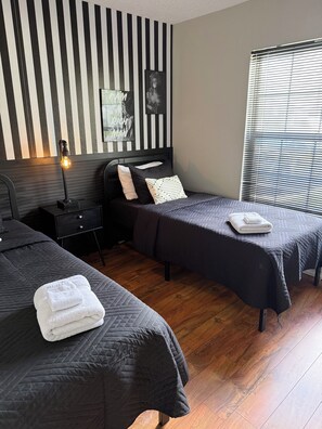 A hotel room with two beds, a nightstand, a lamp, a window with blinds, and a wall with vertical stripes. - Anna Maria Island Getaway | Pool + Game Room | IMG (Bradenton)