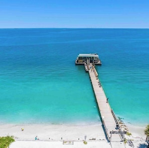 A pier extending into a clear blue sea with a building at the end, a sandy beach, and a clear sky.