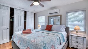Un dormitorio con cama, ventilador de techo, aire acondicionado, mesita de noche con lámpara y ventana con persianas.