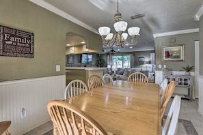A dining area with a wooden table and chairs, a chandelier, and a kitchen in the background.
