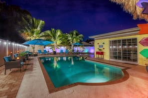 A swimming pool with lounge chairs and umbrellas, surrounded by palm trees and a building with large windows.