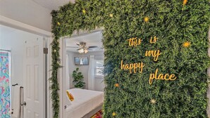 Une pièce avec un lit, un ventilateur de plafond et un mur orné de plantes, sur lequel on peut lire le message : « Voici mon havre de paix. »