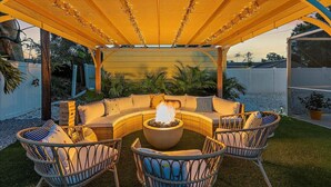 Un espace extérieur circulaire aménagé avec des sièges, doté d’un foyer, entouré de fauteuils et de coussins en osier, et surmonté d’une pergola en bois illuminée de guirlandes lumineuses.