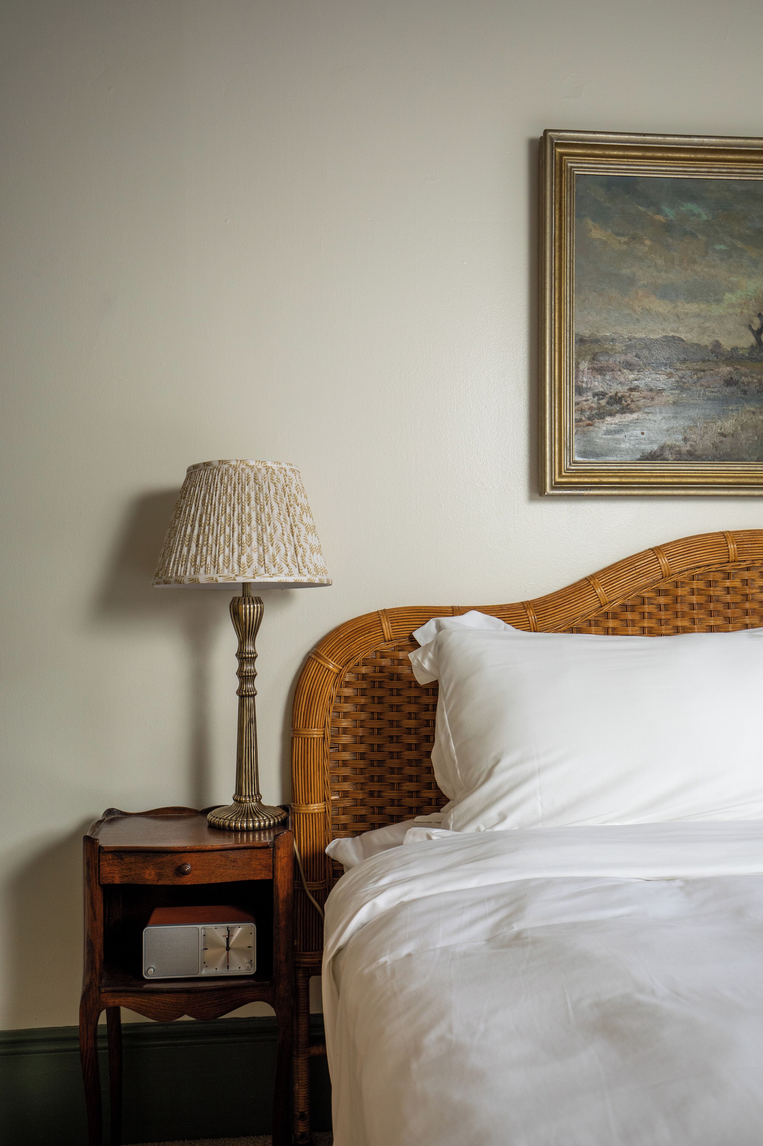 Un lit bien fait, avec du linge de lit blanc, une tête de lit en osier et une table de chevet sur laquelle se trouvent une lampe et une horloge.