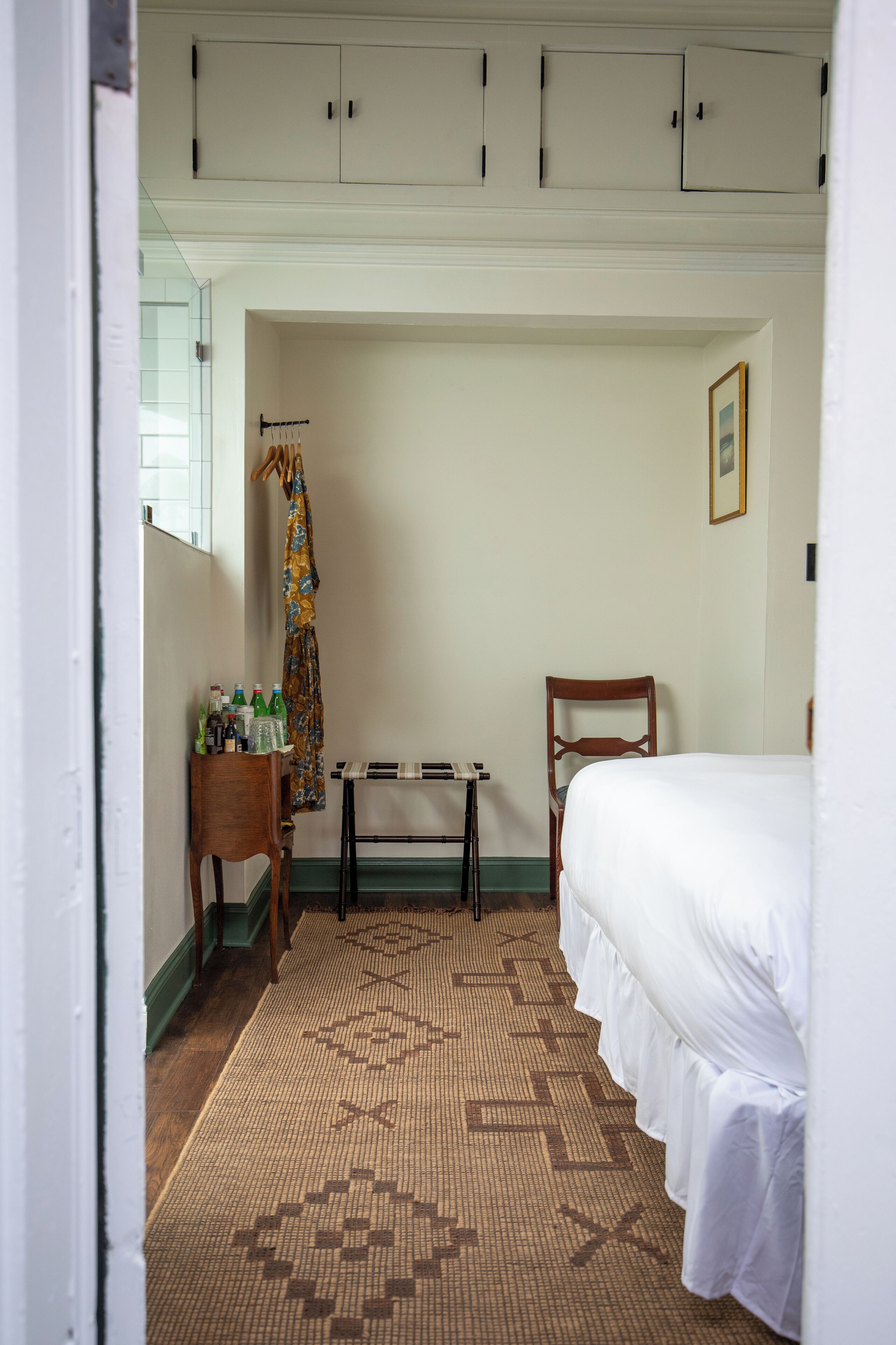 Una camera d'albergo con un letto, una sedia di legno, un tavolino con delle bottiglie e un tappeto decorato.