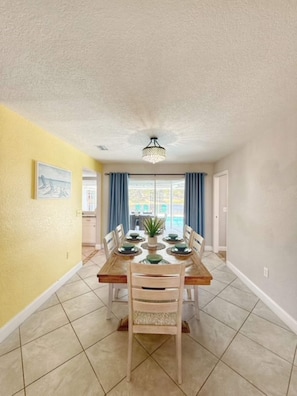 A dining area with a wooden table and chairs, a ceiling light fixture, and a view through a glass door to an outdoor area. - Waterfront Heated Pool | AMI • Siesta • IMG (Sarasota)