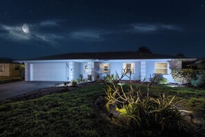A residential house at night with a visible garage, a well-lit front yard, and a clear sky with stars. - Waterfront Heated Pool | AMI • Siesta • IMG (Sarasota)