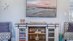 Una sala de estar con chimenea, dos sillones estampados, un adorno con forma de estrella de mar y una pintura de una escena de playa.