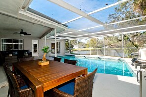 Zona de piscina al aire libre con una mesa y sillas de madera, rodeada por un porche cubierto con ventanas grandes.