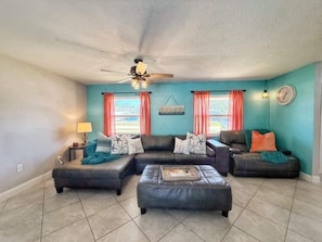A living room with a sectional sofa, an ottoman, a ceiling fan, and a window with curtains. - Waterfront Heated Pool | AMI • Siesta • IMG (Sarasota)