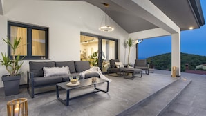 Ein moderner Außenbereich zum Verweilen mit einer Couch, Sesseln und einem Couchtisch. Übertöpfe und ein Blick auf die Landschaft sind ebenfalls vorhanden.