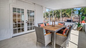 Une terrasse couverte avec une table à manger et des chaises en osier, une porte-fenêtre coulissante et une vue sur une cour arrière agrémentée de palmiers.