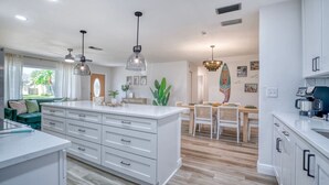 Une cuisine moderne avec des meubles blancs, un îlot central, des luminaires suspendus et un coin repas comprenant une table et des chaises en bois.