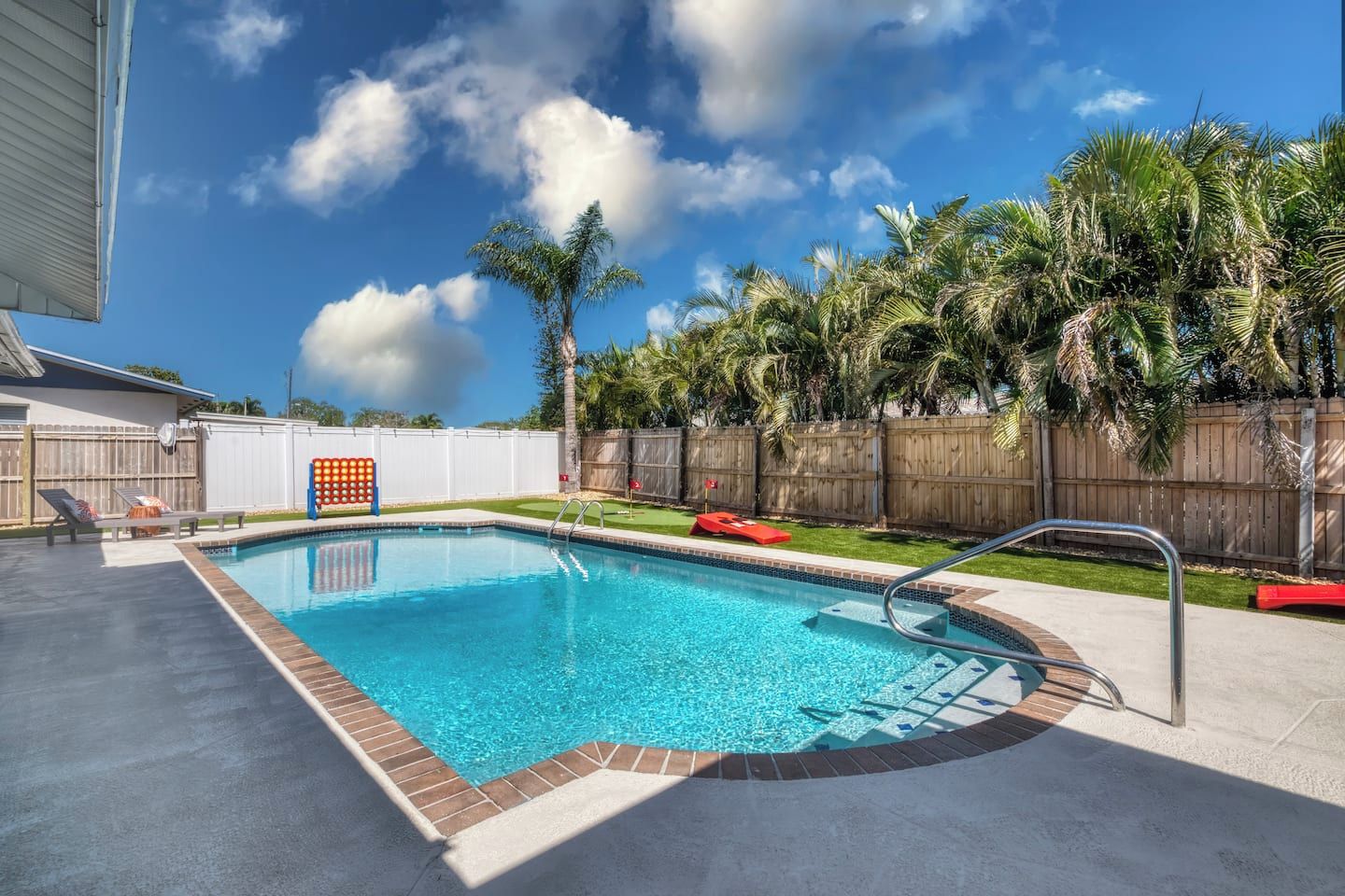 A rectangular swimming pool with a wooden deck and metal ladder, surrounded by a wooden fence and palm trees.