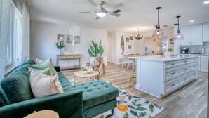 Un vaste salon avec un canapé vert, un coin repas, une cuisine dotée de meubles blancs et un ventilateur de plafond.