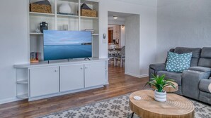 Una sala de estar moderna con un televisor de pantalla plana sobre un mueble blanco, un sofá gris con cojines estampados y una mesa de centro redonda de madera con una planta.