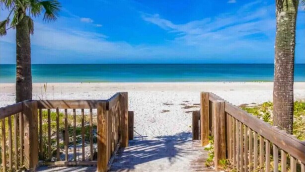 Un sentiero di legno conduce a una spiaggia con palme e un cielo azzurro terso.